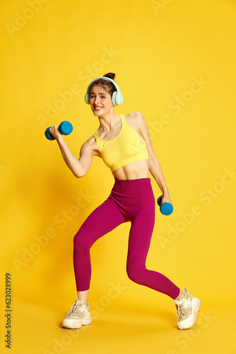 Full-length image of young slim woman in sportswear and headphones training with heavy dumbbells against yellow studio background. Concept of sport, fitness, body care, fashion, youth, lifestyle, ad