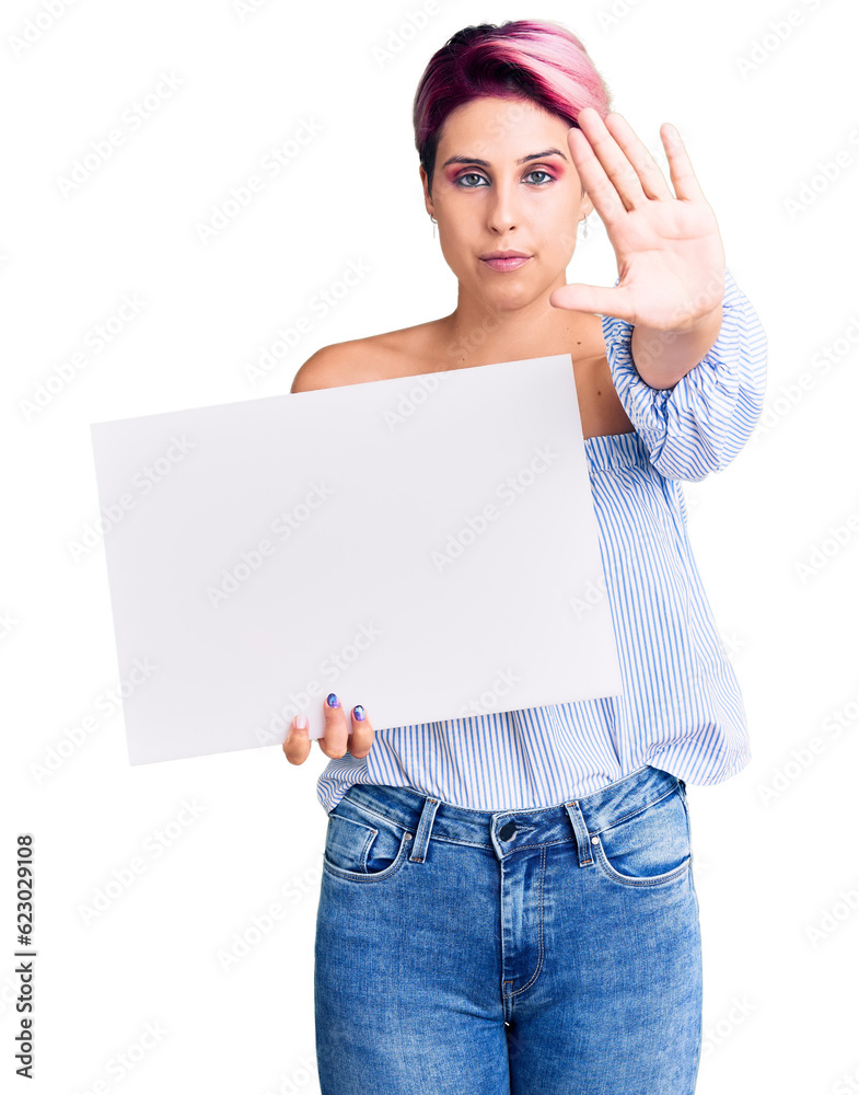 Young beautiful woman with pink hair holding cardboard banner with blank space with open hand doing stop sign with serious and confident expression, defense gesture Young beautiful woman with pink hair holding cardboard banner with blank space with open hand doing stop sign with serious and confident expression, defense gesture