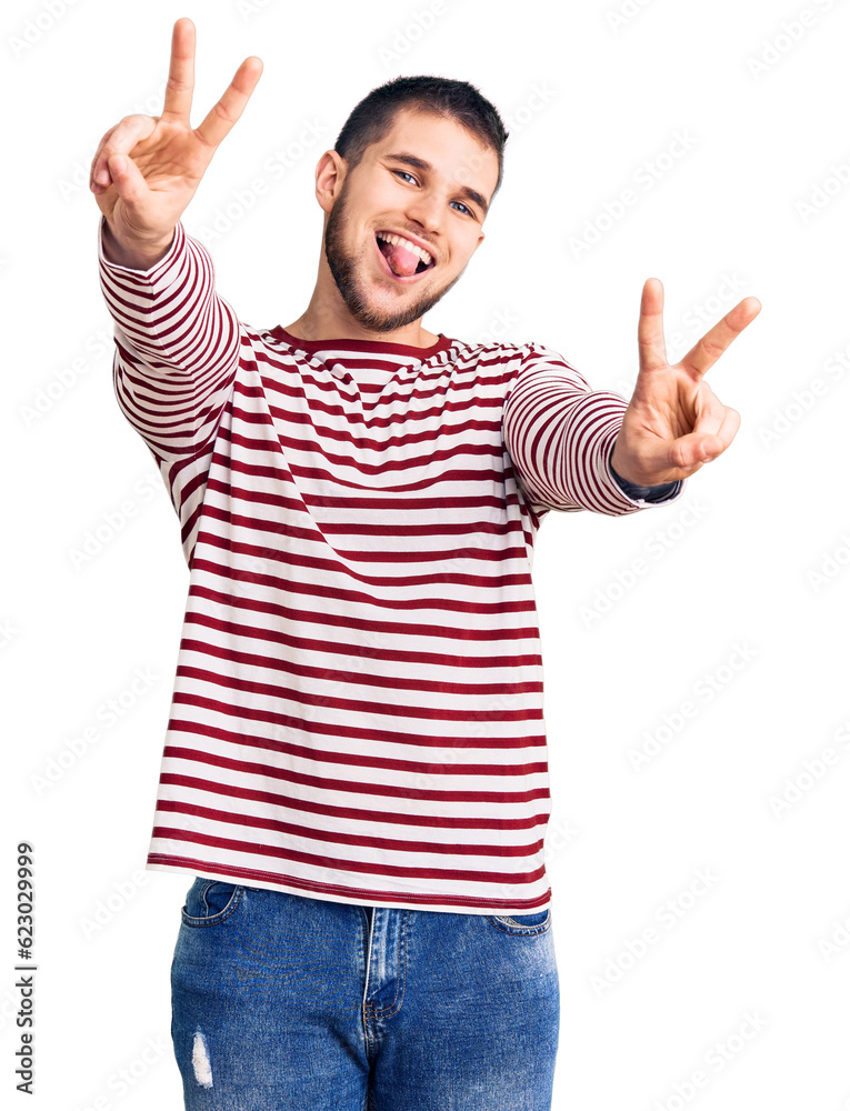 Young handsome man wearing striped sweater smiling with tongue out showing fingers of both hands doing victory sign. number two. Young handsome man wearing striped sweater smiling with tongue out showing fingers of both hands doing victory sign. number two.