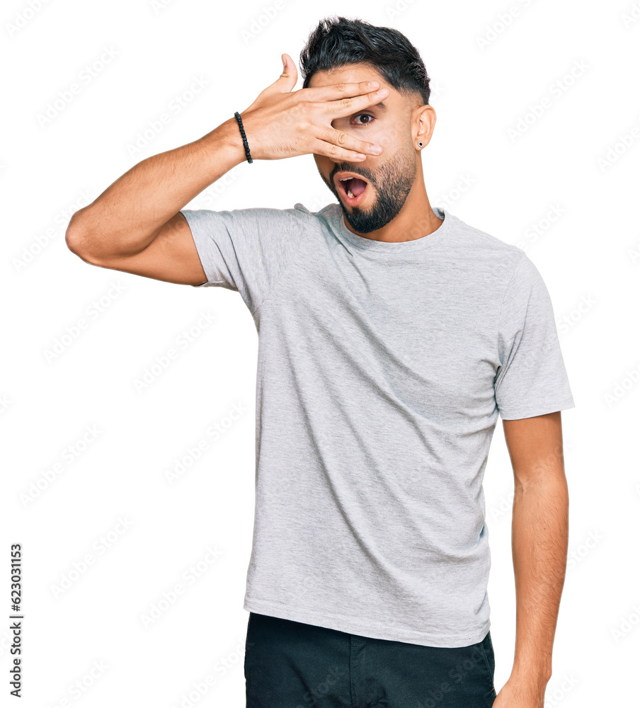 Young man with beard wearing casual grey tshirt peeking in shock covering face and eyes with hand, looking through fingers with embarrassed expression. Young man with beard wearing casual grey tshirt peeking in shock covering face and eyes with hand, looking through fingers with embarrassed expression.