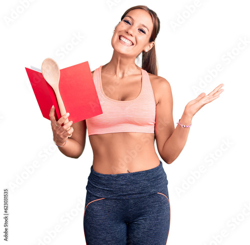 Young beautiful hispanic woman wearing gym clothes reading cooking recipe book holding spoon celebrating victory with happy smile and winner expression with raised hands