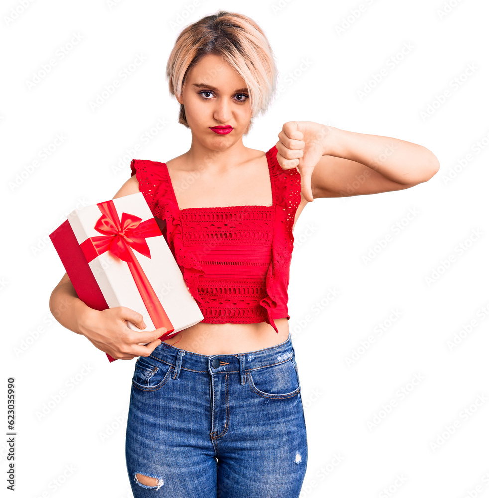 Young beautiful blonde woman holding gift with angry face, negative sign showing dislike with thumbs down, rejection concept Young beautiful blonde woman holding gift with angry face, negative sign showing dislike with thumbs down, rejection concept