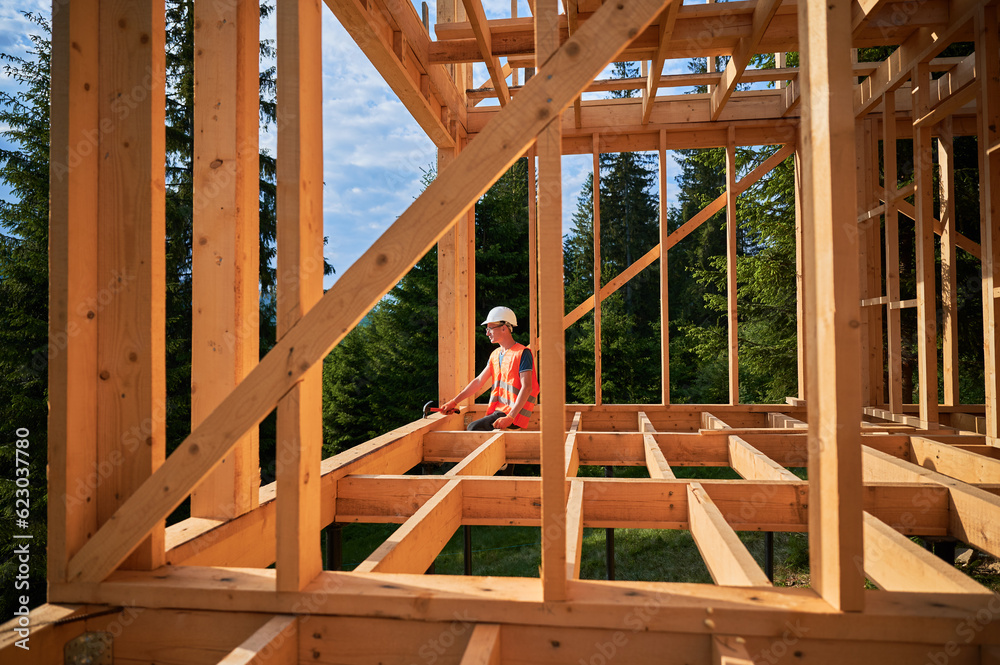Carpenter constructing wooden frame two-story house near the forest. Man in glasses holding hammer, dressed in protective helmet and orange safety vest. Concept of ecological modern construction.