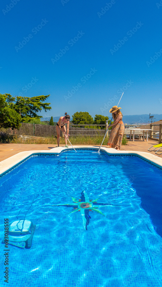 Two lesbian couple friends cleaning a swimming pool together dressed in summer dress and bikini with pool cleaning poles with basket and brush with clear blue sky in the background on a sunny day