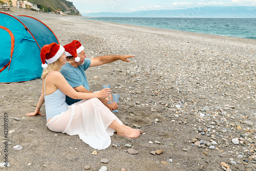 Middle aged couple wearing santa hats having christmas picnic on the beach. Bearded man and pretty blond woman drinking sparkling wine and having fun looking the sea.