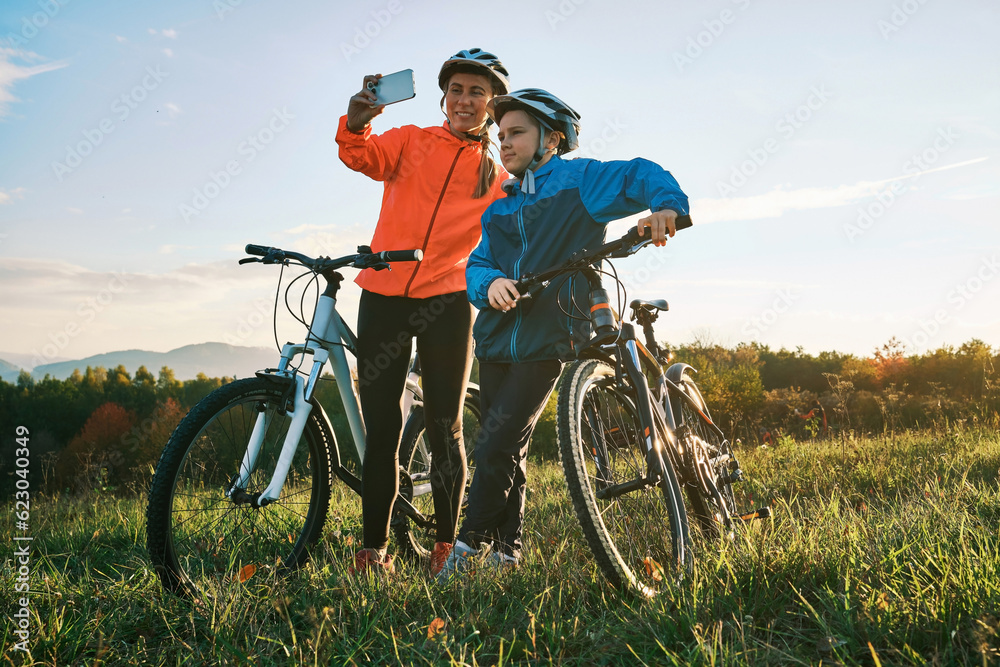 Mother and son ride a bike in park on green meadow in autumn day at sunset time. Family weekend. Woman traveler using smartphone, taking photo

