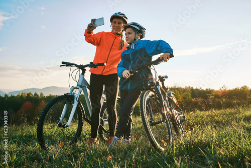 Mother and son ride a bike in park on green meadow in autumn day at sunset time. Family weekend. Woman traveler using smartphone, taking photo 