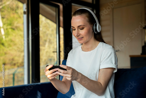Beautiful woman is playing a mobile game and wearing wireless headphones while sitting on a blue sofa in the living room. Playing the game on a smartphone, you win the moment of victory