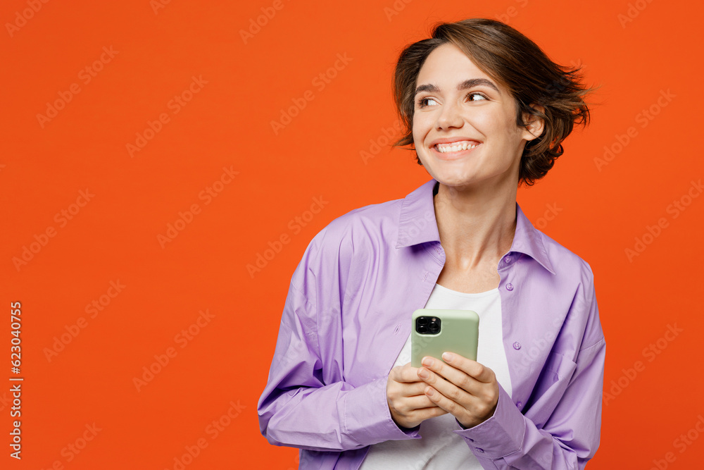 © ViDi Studio - Young caucasian woman wear purple shirt white t-shirt casual clothes hold in hand use mobile cell phone look aside on workspace isolated on plain orange background studio portrait. Lifestyle concept.