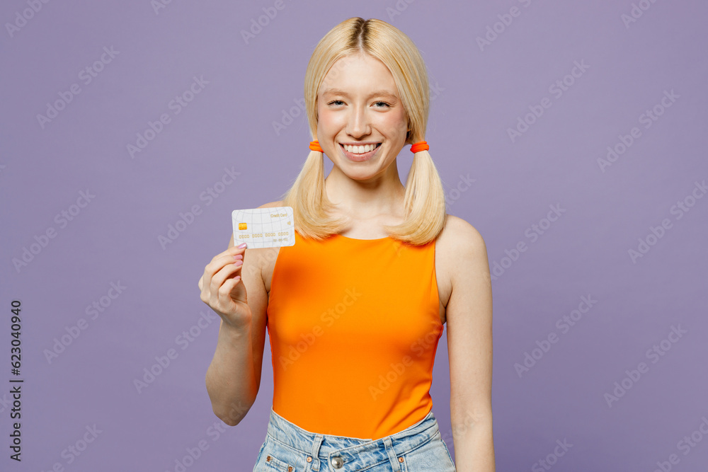 Young cheerful smiling blonde woman wear orange tank shirt casual clothes hold in hand mock up of credit bank card isolated on plain pastel light purple background studio portrait. Lifestyle concept. Young cheerful smiling blonde woman wear orange tank shirt casual clothes hold in hand mock up of credit bank card isolated on plain pastel light purple background studio portrait. Lifestyle concept.
