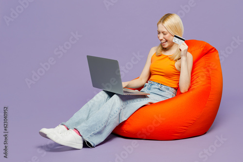 Full body young blonde woman wear orange tank shirt casual clothes sit in bag chair using laptop pc computer hold credit bank card shopping online order delivery isolated on plain purple background.