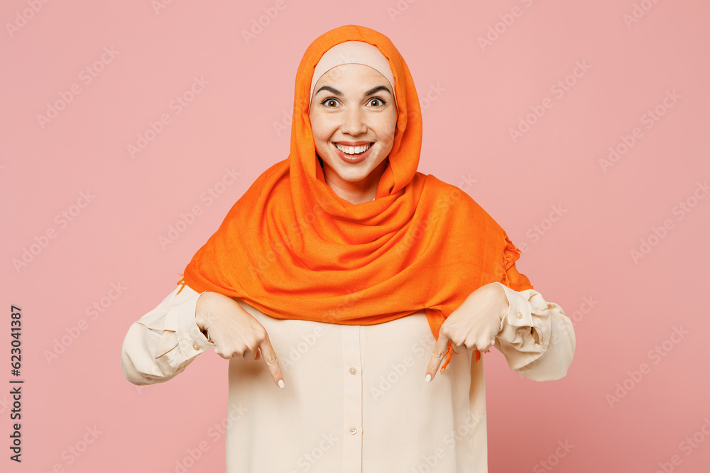 Fototapeta premium Young arabian asian muslim woman wear orange abaya hijab point index finger down on area workspace isolated on plain light pink background studio portrait. Uae middle eastern islam religious concept.