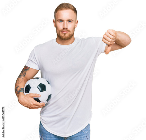 Young irish man holding soccer ball with angry face, negative sign showing dislike with thumbs down, rejection concept