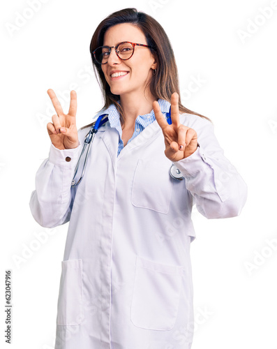 Young beautiful woman wearing doctor stethoscope and glasses smiling looking to the camera showing fingers doing victory sign. number two.