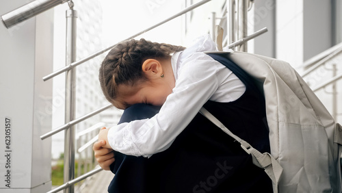 Brown-haired schoolgirl sitting on empty stairs cries remembering bullying moments. Girl with long braids afraid to attend school and see bullies photo