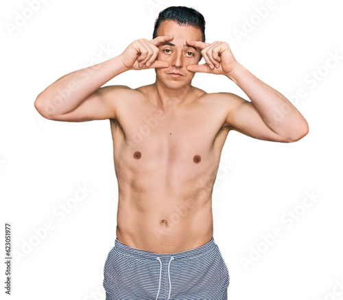 Handsome young man wearing swimwear shirtless trying to open eyes with fingers, sleepy and tired for morning fatigue