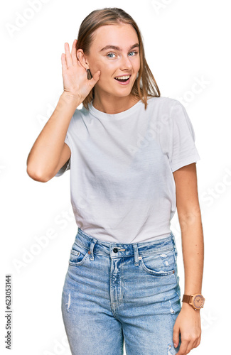 Beautiful young blonde woman wearing casual white t shirt smiling with hand over ear listening an hearing to rumor or gossip. deafness concept.