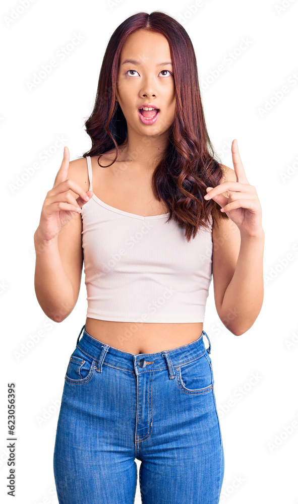 Young beautiful chinese girl wearing casual clothes amazed and surprised looking up and pointing with fingers and raised arms. Young beautiful chinese girl wearing casual clothes amazed and surprised looking up and pointing with fingers and raised arms.