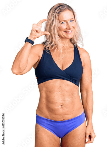 Middle age fit blonde woman wearing bikini smiling and confident gesturing with hand doing small size sign with fingers looking and the camera. measure concept. © Krakenimages.com