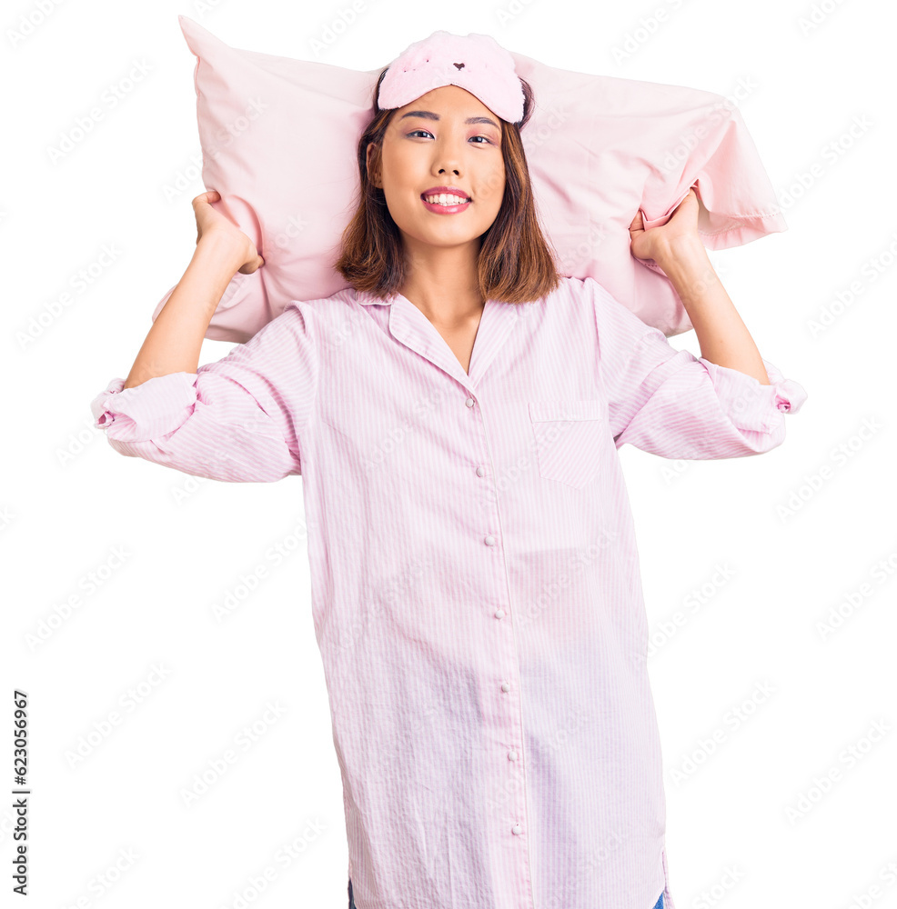 Young beautiful chinese girl wearing sleep mask and pajama holding pillow looking positive and happy standing and smiling with a confident smile showing teeth Young beautiful chinese girl wearing sleep mask and pajama holding pillow looking positive and happy standing and smiling with a confident smile showing teeth