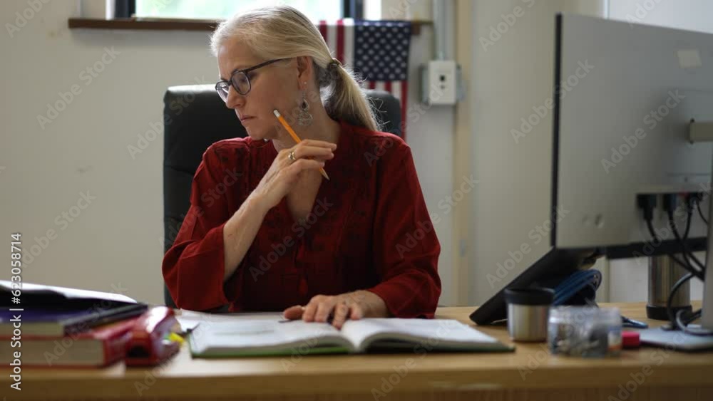 Closeup of happy woman teacher sitting at desk in empty school classroom desk grading papers working on assignments. US American flag in the background.
