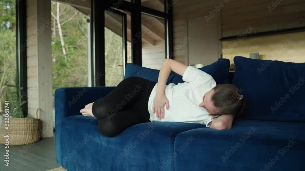 Sad young woman dressed in casual clothes, suffering from menstrual pains, feeling nausea in her stomach, holding her stomach, experiencing abdominal cramps during menstruation and lying at home on a Sad young woman dressed in casual clothes, suffering from menstrual pains, feeling nausea in her stomach, holding her stomach, experiencing abdominal cramps during menstruation and lying at home on a