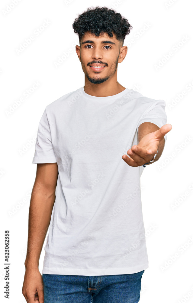 Young arab man wearing casual white t shirt smiling cheerful offering palm hand giving assistance and acceptance. Young arab man wearing casual white t shirt smiling cheerful offering palm hand giving assistance and acceptance.