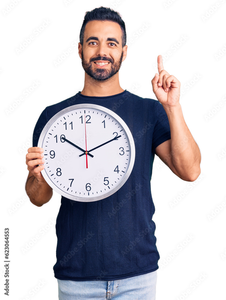 Young hispanic man holding big clock surprised with an idea or question pointing finger with happy face, number one Young hispanic man holding big clock surprised with an idea or question pointing finger with happy face, number one
