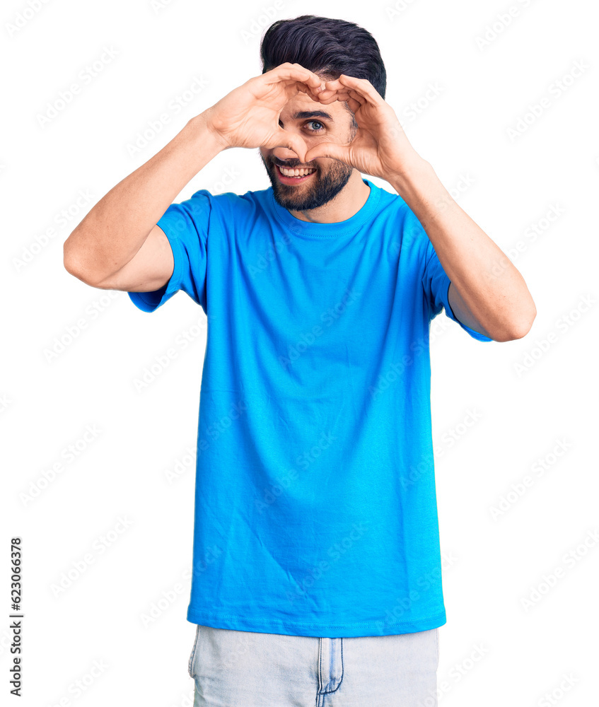 Young handsome man with beard wearing casual t-shirt doing heart shape with hand and fingers smiling looking through sign Young handsome man with beard wearing casual t-shirt doing heart shape with hand and fingers smiling looking through sign