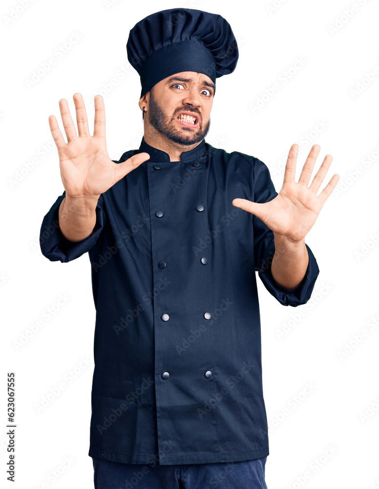 Young hispanic man wearing cooker uniform afraid and terrified with fear expression stop gesture with hands, shouting in shock. panic concept.