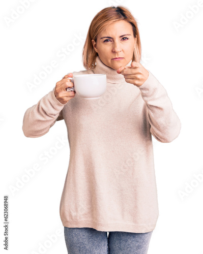 Young blonde woman holding coffee pointing with finger to the camera and to you, confident gesture looking serious