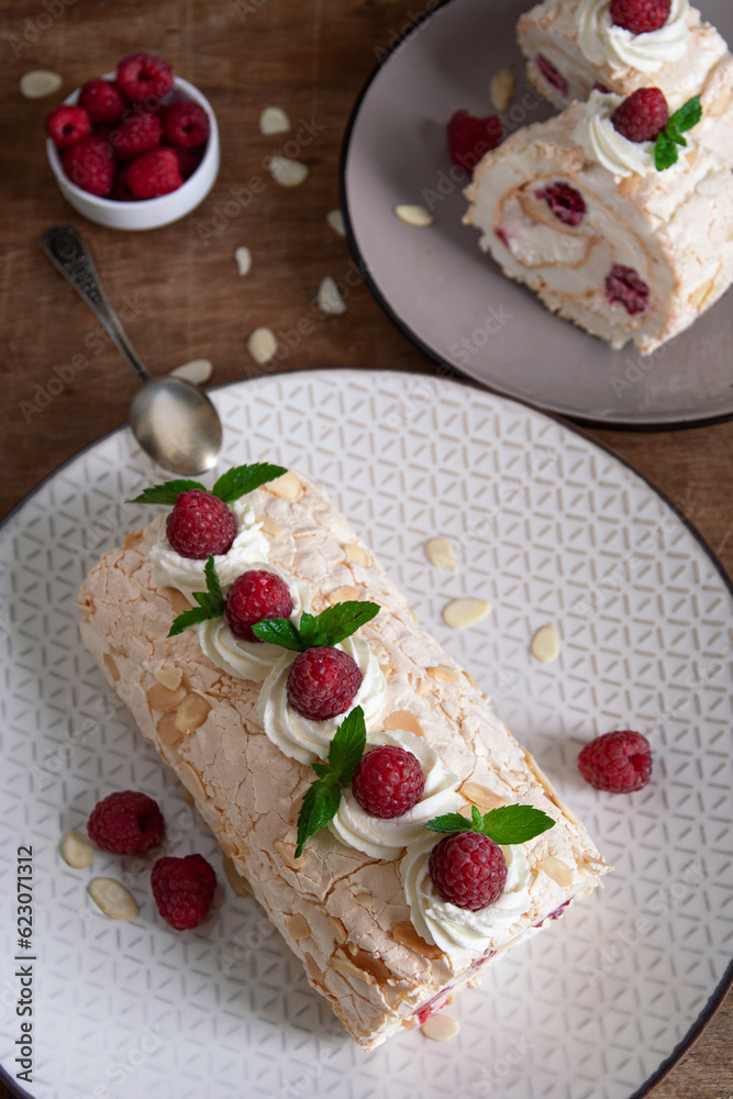 Top view of a meringue roll with raspberries and almond petals, decorated with cream, raspberries and mint leaves, on a wooden background. Vertical Top view of a meringue roll with raspberries and almond petals, decorated with cream, raspberries and mint leaves, on a wooden background. Vertical