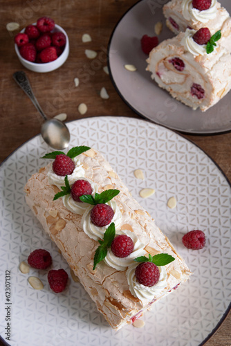 Top view of a meringue roll with raspberries and almond petals, decorated with cream, raspberries and mint leaves, on a wooden background. Vertical