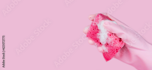 A women's hand in pink rubber glove holds a cleaning sponge with soapy foam on a pink background. Copy space. Banner. Selective focus.
