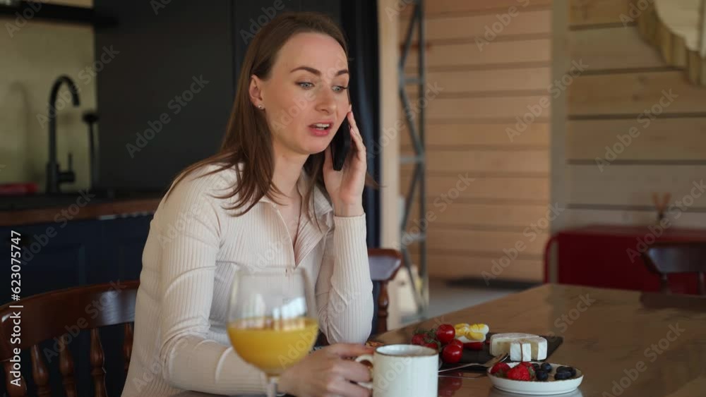 Cheerful young woman talking on the phone in the kitchen sitting at the dining table. Cheerful young woman talking on the phone in the kitchen sitting at the dining table.