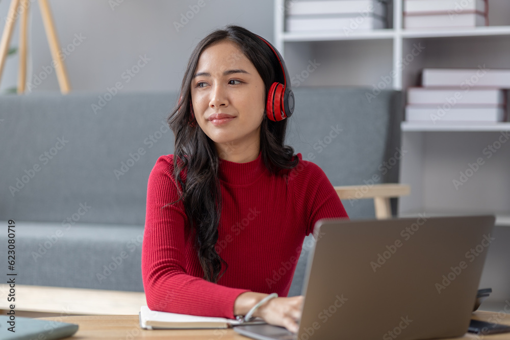 Asian beautiful woman sits in living room, she is a college student listening to music and playing social media on laptop for relaxing, studying from home, studying online. University student concept. Asian beautiful woman sits in living room, she is a college student listening to music and playing social media on laptop for relaxing, studying from home, studying online. University student concept.