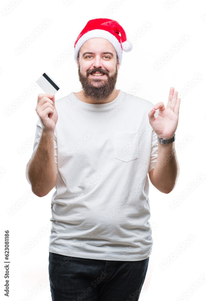 Young caucasian man wearing christmas hat holding credit card over isolated background doing ok sign with fingers, excellent symbol