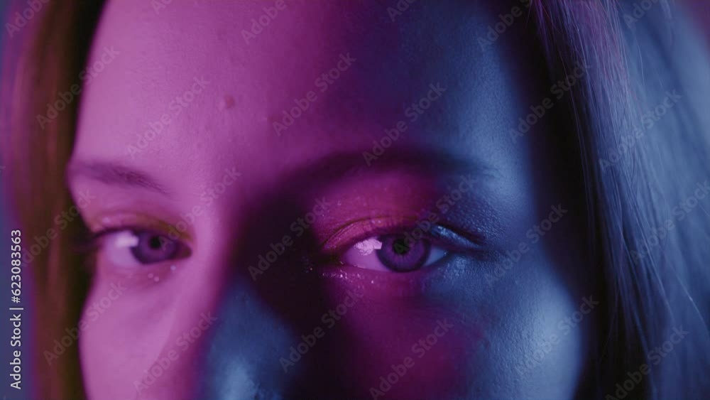 Portrait of a close-up opening eyes of a beautiful girl in a slow motion. Attractive millennial woman in the studio, neon blue pink light. Professional female model. Beauty fashion with glitter