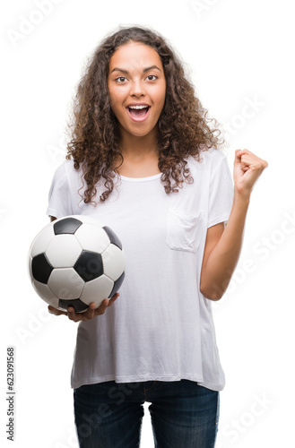 Young hispanic woman holding soccer football ball screaming proud and celebrating victory and success very excited, cheering emotion