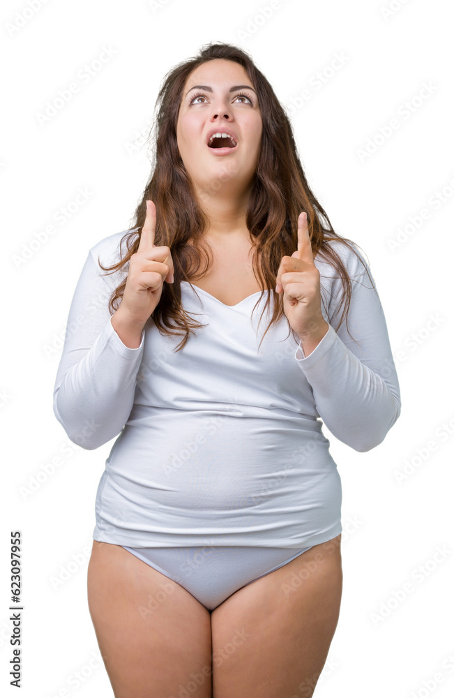Beautiful plus size young overwight woman wearing white underwear over isolated background amazed and surprised looking up and pointing with fingers and raised arms. Beautiful plus size young overwight woman wearing white underwear over isolated background amazed and surprised looking up and pointing with fingers and raised arms.