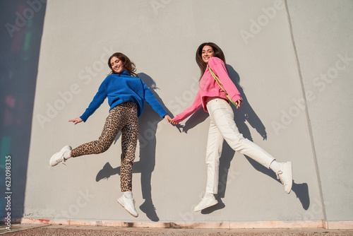 Lovely besties holding hands and looking at the camera while jumping near the wall photo