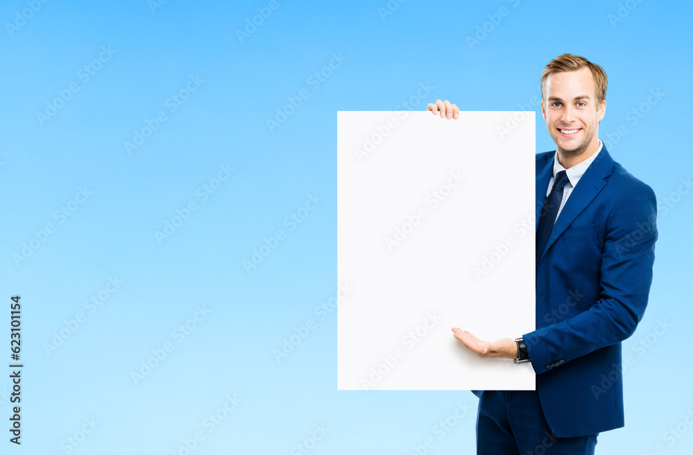 Businessman showing white empty paper sign board with copyspace area, over blue color background. Portrait of happy smiling confident man in business studio concept. Businessman showing white empty paper sign board with copyspace area, over blue color background. Portrait of happy smiling confident man in business studio concept.