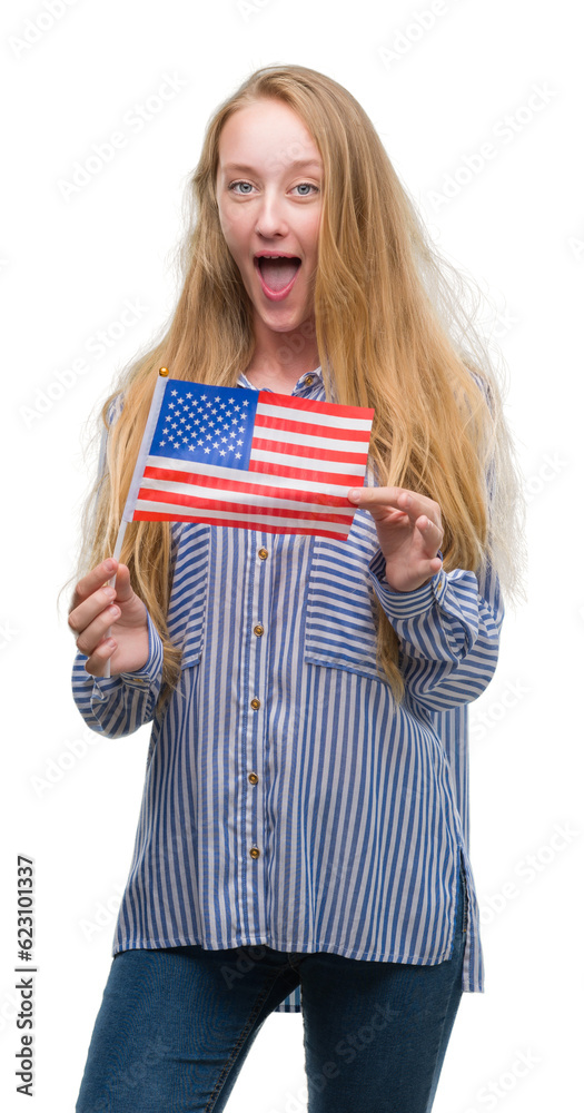 Blonde teenager woman holding flag of United States of America scared in shock with a surprise face, afraid and excited with fear expression Blonde teenager woman holding flag of United States of America scared in shock with a surprise face, afraid and excited with fear expression