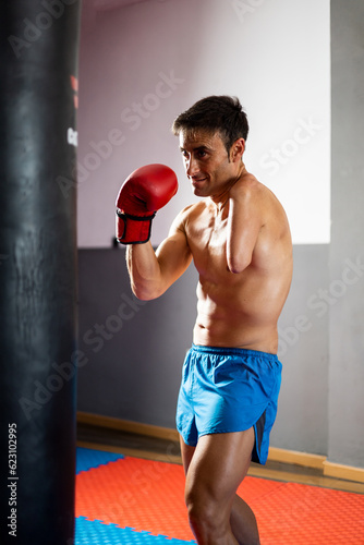 Vertical photo of a shirtless boxer with an amputated arm poses in front of a punching bag inside a gym. Boxing concept, disabled boxer, an adult in a boxing glove with an amputated arm.Improvement.