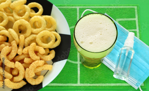 Puffed corn snacks on soccer ball plate, focus on top of beer glass, green football field dish mat. Blue medical mask and sanitizer, watching match broadcast with safety during covid pandemic.