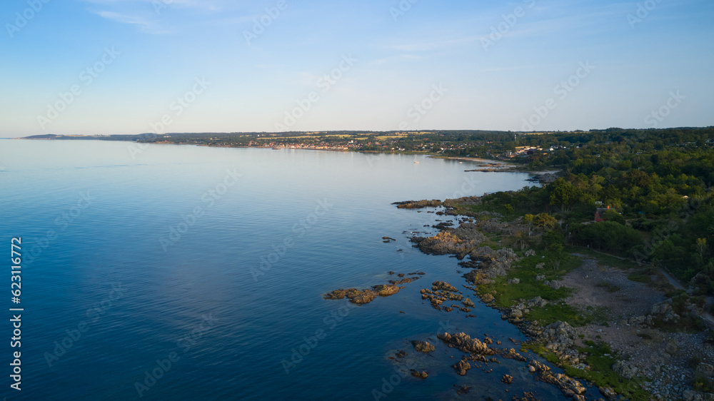 Blick von oben auf die Granitküste in Richtung der dänischen Ortschaften Tejn und Sandkaas auf der Ostsee-Insel Bornholm an einem sonnigen Abend vor blauem Himmel und geringer Bewölkung