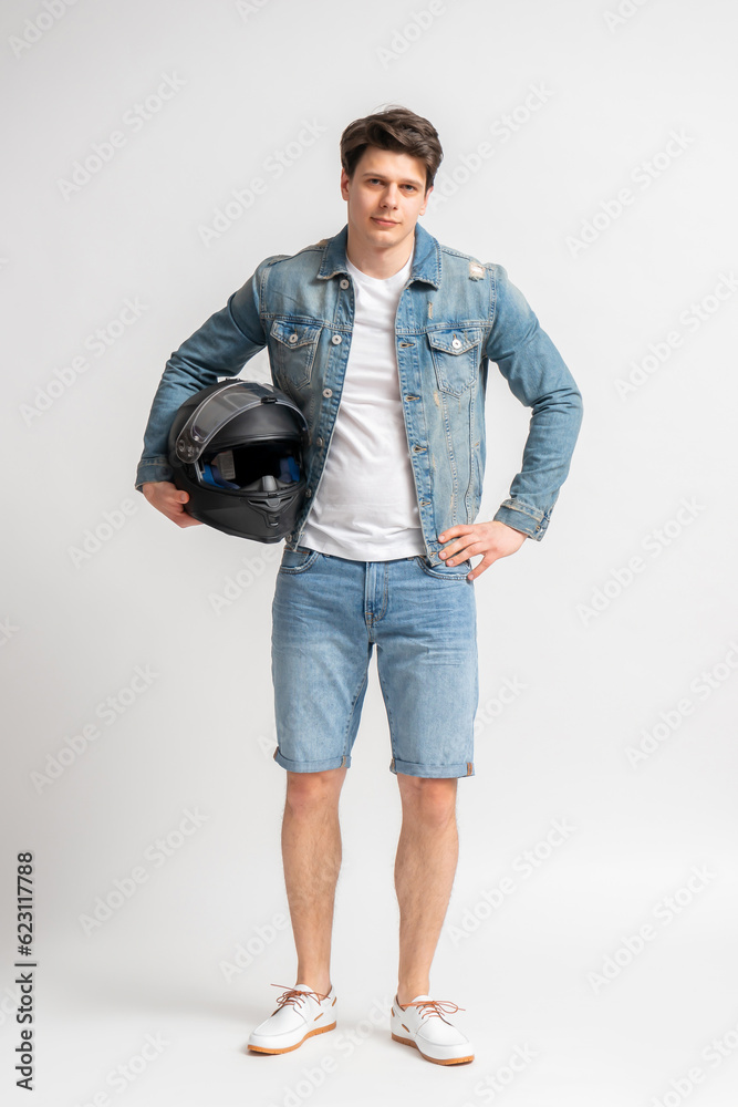 A man in a casual denim jacket and shorts holds a protective motorcycle helmet. A man in a casual denim jacket and shorts holds a protective motorcycle helmet.