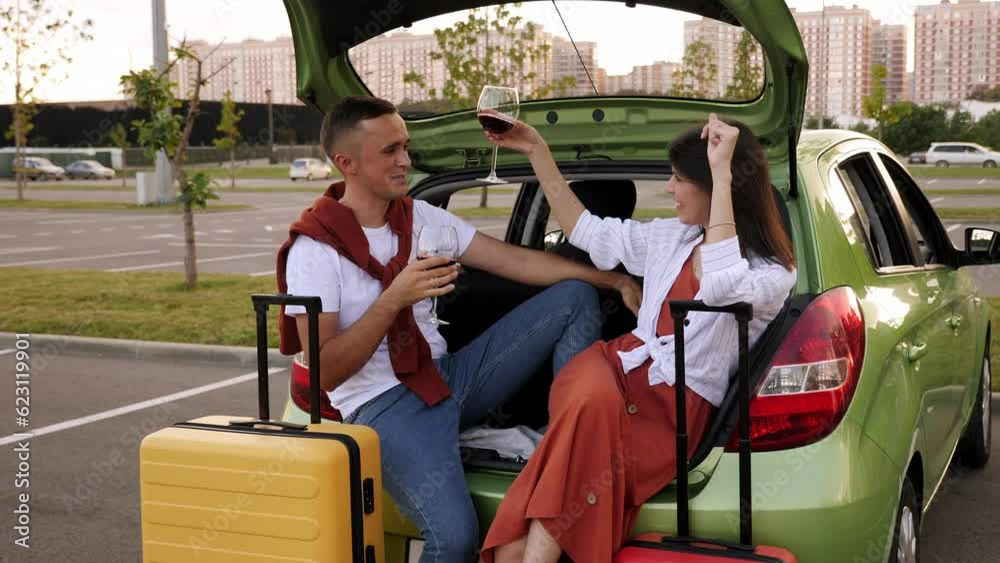 A young couple clink glasses and drink wine sitting in an open trunk while traveling by car. A young couple clink glasses and drink wine sitting in an open trunk while traveling by car.