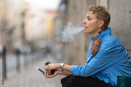 Adult stylish woman sitting on the european street and smoking an electronic cigarette