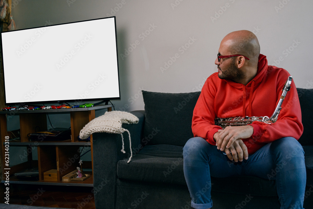 young latino musician man sitting with his saxophone at home turned to watch the tv screen.